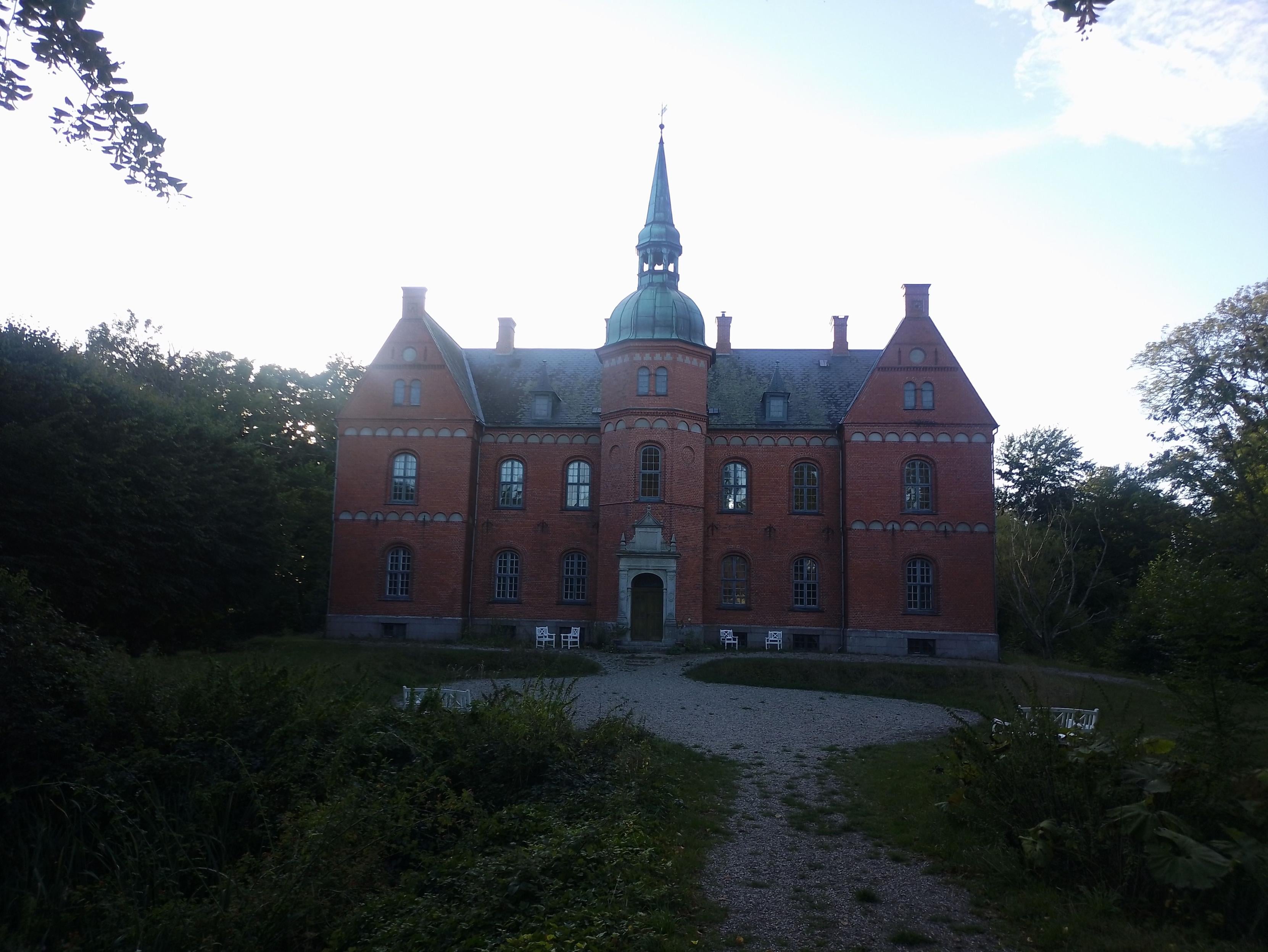 A small red brick castle seen from the bridge across the moat, towards the main entrance. The castle has one central greenish cobber clad tower in front of a two-story wing with two cross-wings that have pointed gables.