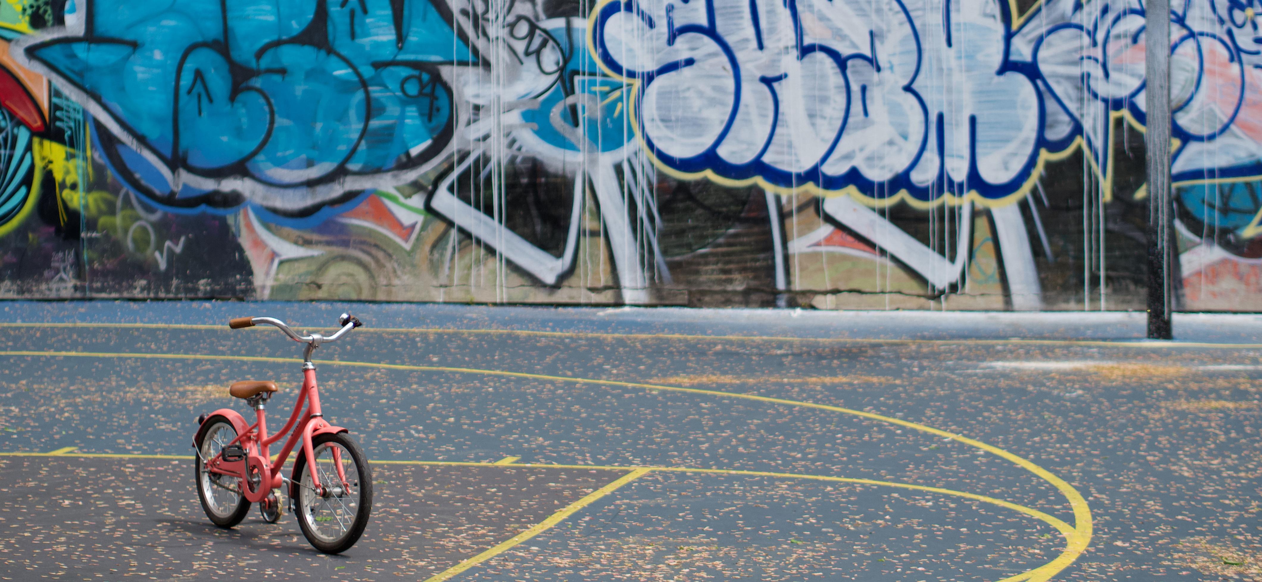 A small orangish-red kids bicycle sits in a basketball court, before a colorful graffiti covered background.